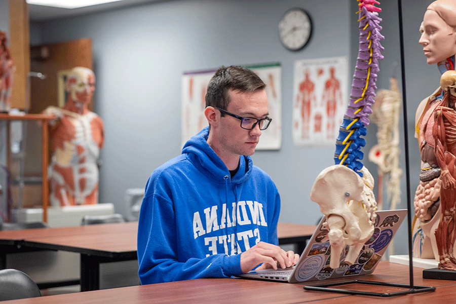 A man with short, dark brown hair and glasses, types on his computer in an anatomy lab. He wears a blue Indiana State hoodie.  There are spine and human body models around him in the lab.