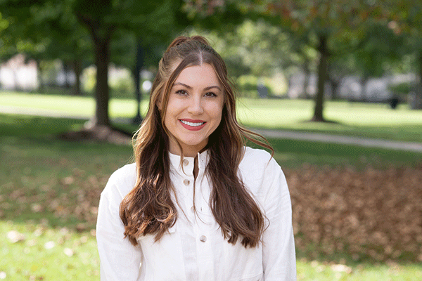 A white female student with wavy brown hair poses outside, wearing a white button-up shirt. Trees and fallen leaves are visible behind her. 